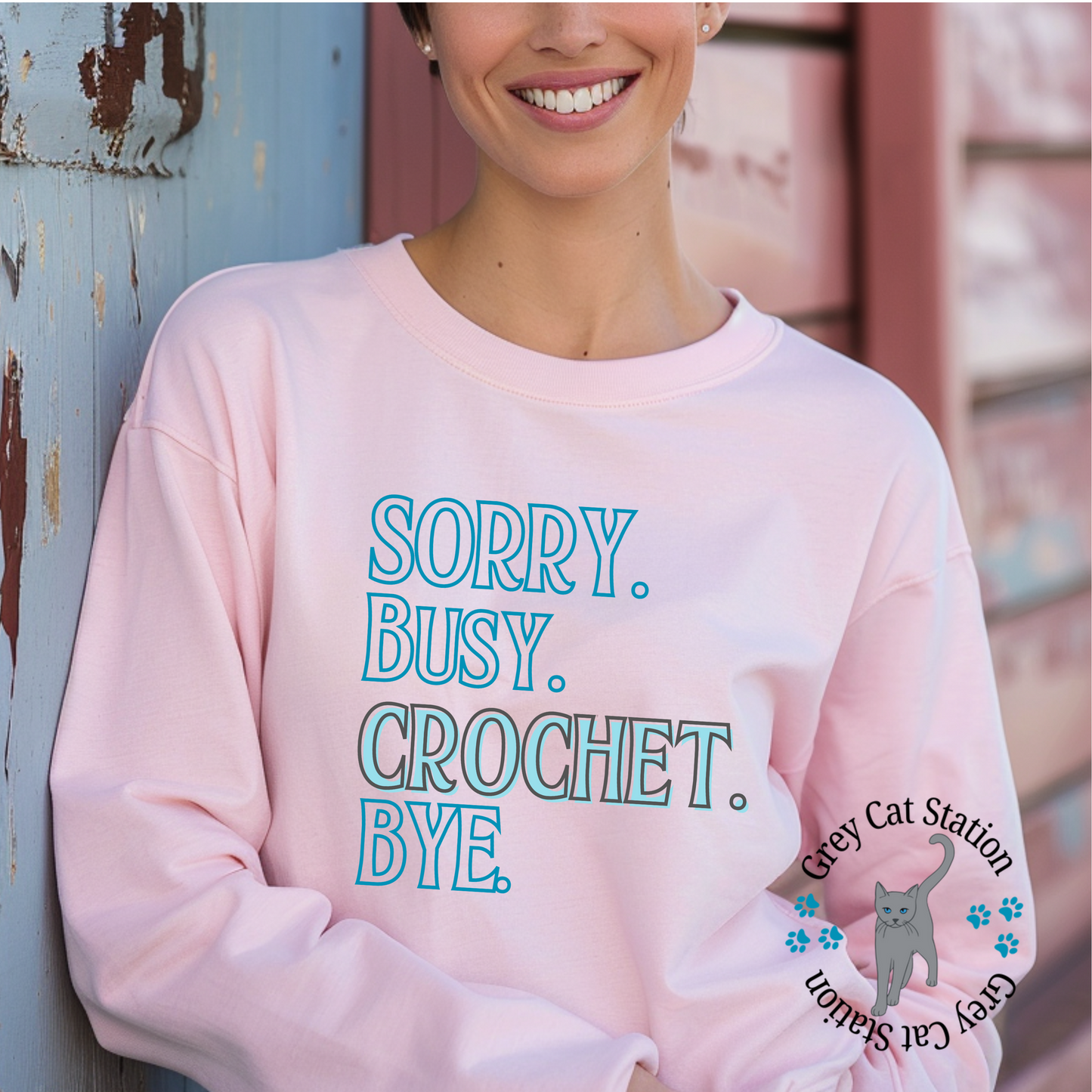 Person wearing a pink sweatshirt with text 'Sorry. Busy. Crochet. Bye.' and a logo on a rustic wooden background.